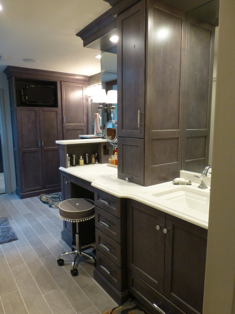 Dark bathroom cabinetry with tall storage, a seated dressing area, drawers and a sink along a light countertop.