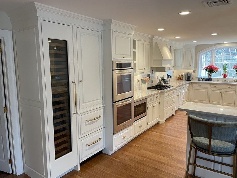 White kitchen cabinetry surrounding a tall glass-front appliance and stacked ovens beside an island.