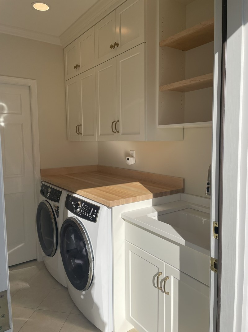 White laundry room cabinets above a washer and dryer, with a wood countertop and adjacent sink.