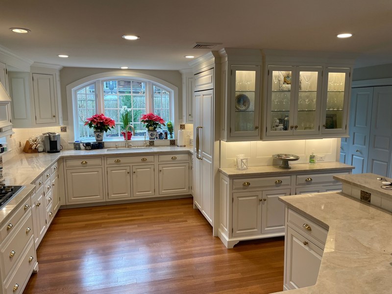 White kitchen cabinets with illuminated glass-front upper doors, a window over the sink and wood flooring.