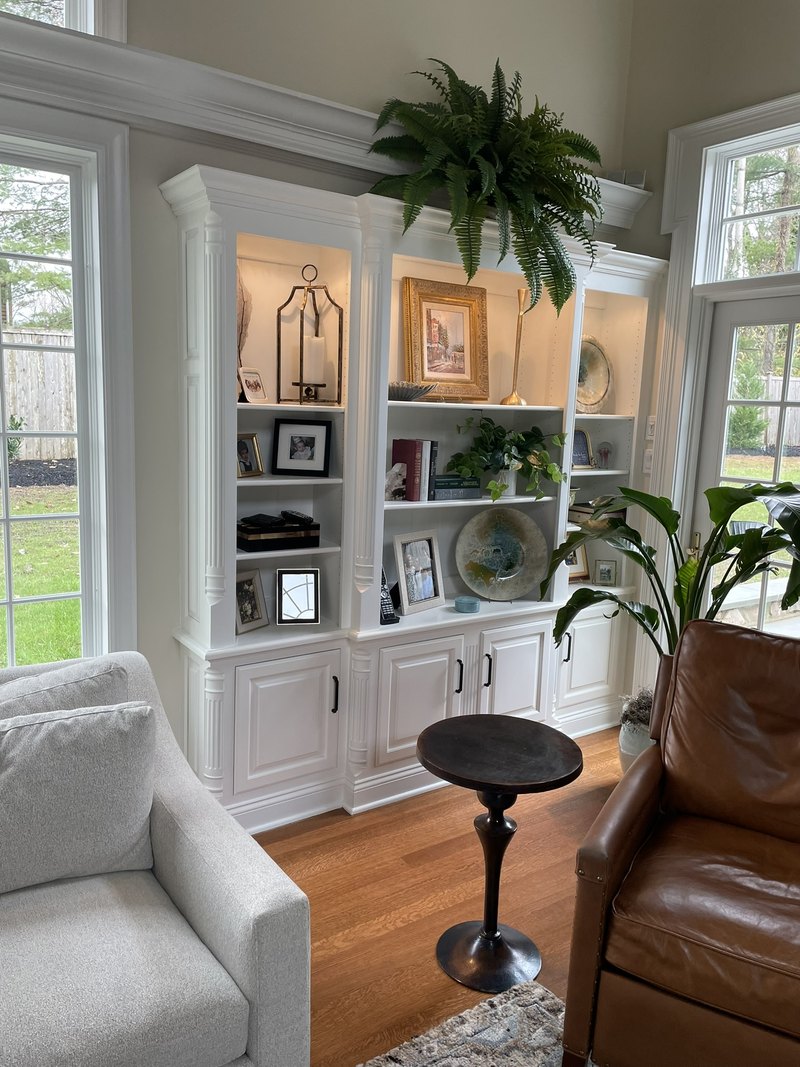 White built-in display shelves and lower cupboards between windows in a furnished living room.