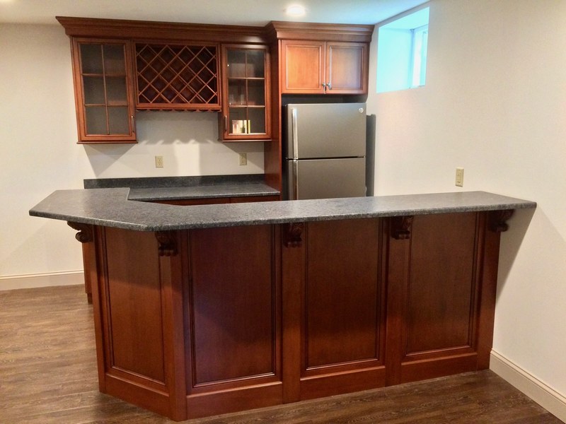 Wood home bar with paneled front, dark countertop, upper wine rack and a refrigerator.