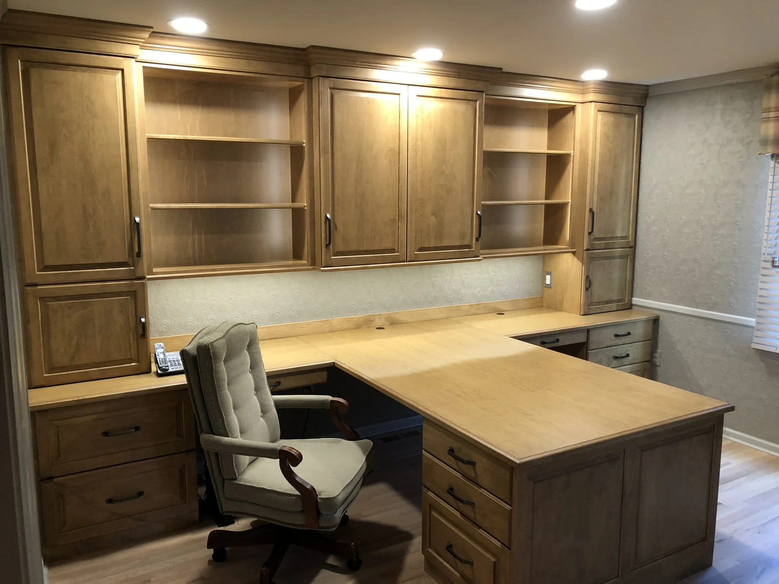 Wood home office cabinetry with an L-shaped desk, drawers, upper cabinets and illuminated open shelves.