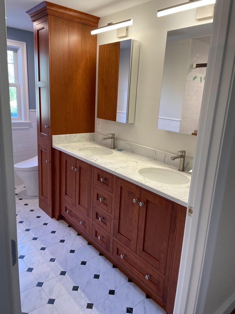 Warm wood double bathroom vanity with center drawers and a matching tall linen cabinet beside the sinks.